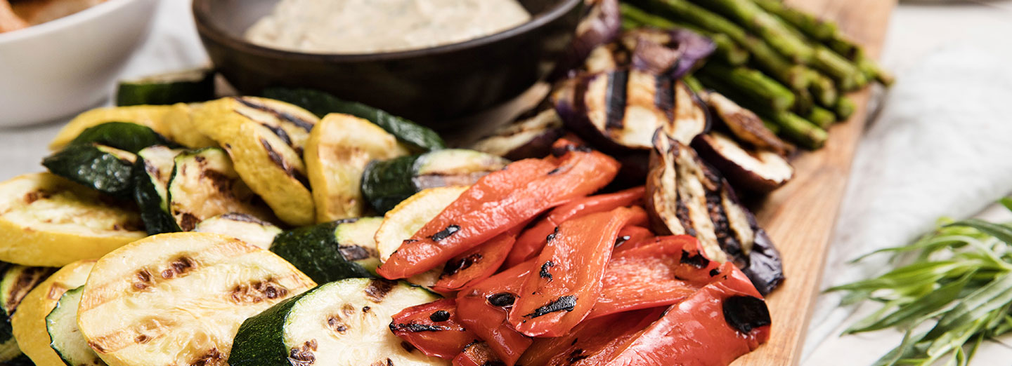 grilled vegetables arranged on cutting board surrounded by herbs with garlic mustard sauce on side