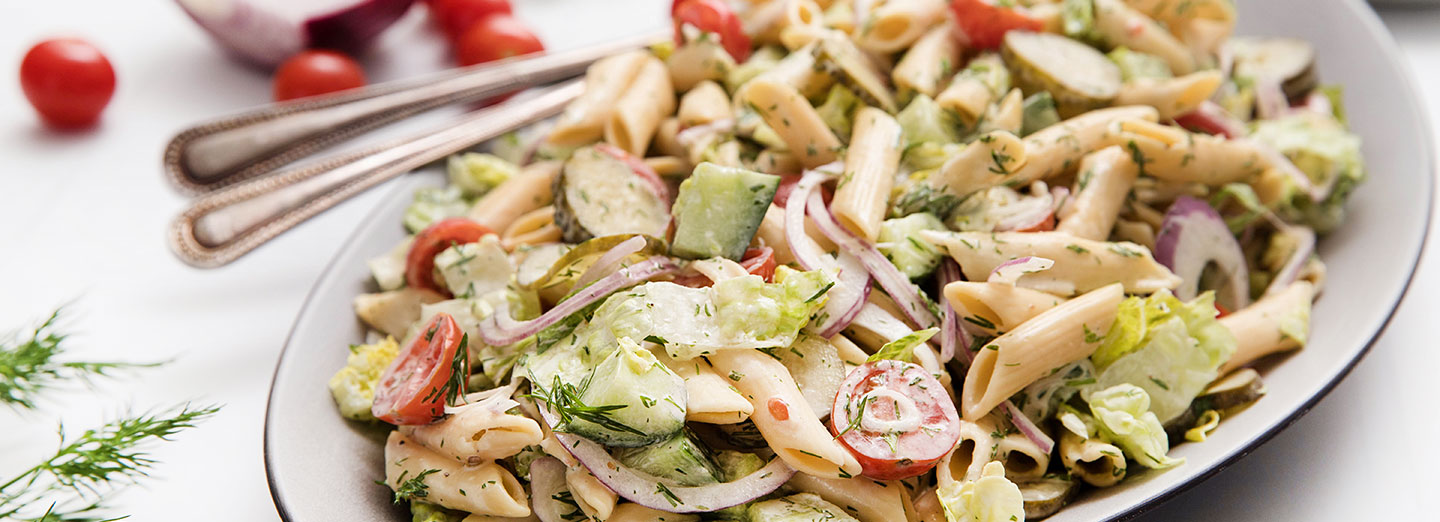 pasta salad topped with pickles, tomatoes, onions and dill on a plate on a white table